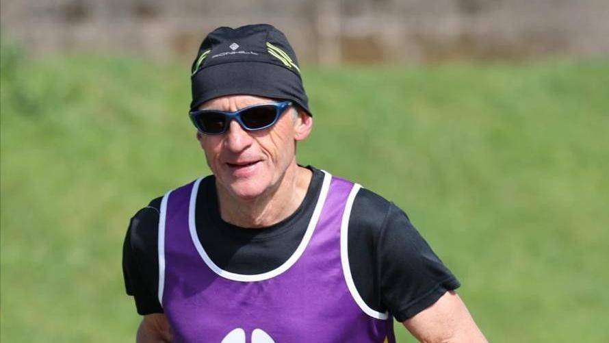 A man wearing a black hat and T-shirt, sunglasses and a purple bib, running with grassland in the background.