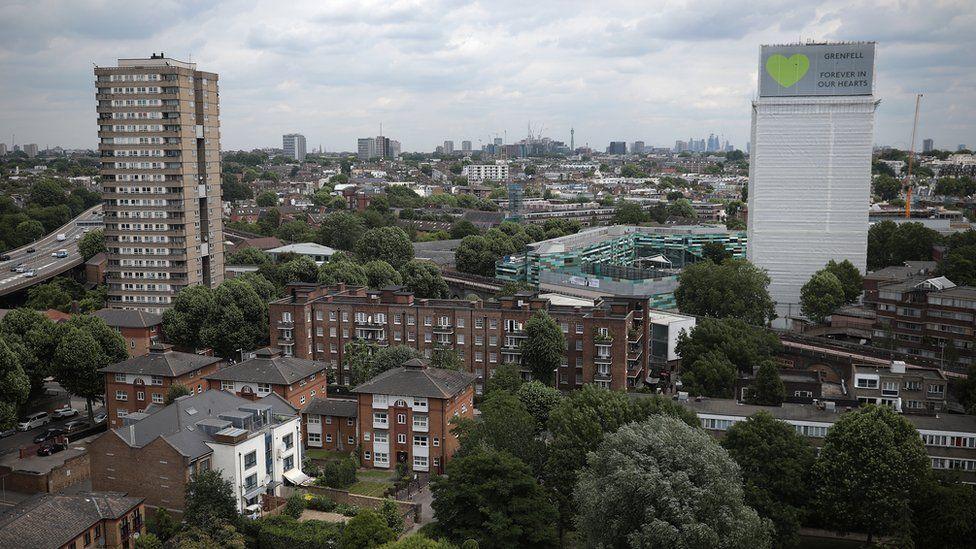 Grenfell Tower wrapped in white sheeting, with a green heart and the words “Grenfell Forever in Our Hearts” at the top, overlooking surrounding homes and buildings in west London.