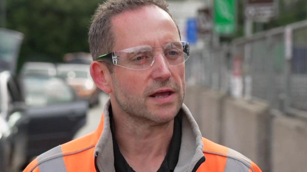 Daniel has short brown hair and is wearing protective glasses. He is also wearing an orange high vis jacket.