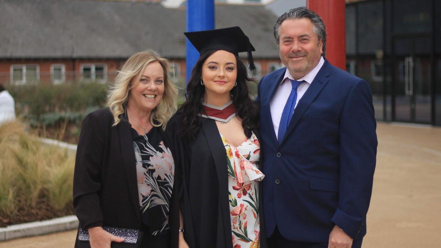 Lauren stands with her parents at her graduation. She is wearing a graduates cap and gown over a white floral dress. Her mum stand next to her on the left wearing a black blazer and trousers and floral blouse. Lauren's dad stands on her right, he is wearing a blue suit with a pink shirt and a blue tie.