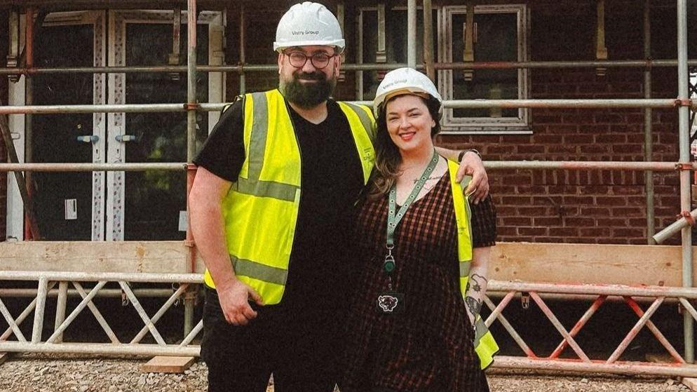 Charles and Georgina pose on a muddy building site in front of a partially completed brick house surrounded by scaffolding. Both wear safety helmets, high-visibility vests and sturdy boots. Stacks of timber lie in the foreground, while the unfinished structure, exposed windows and metal scaffold framework fill the background, illustrating the progress of the construction project.