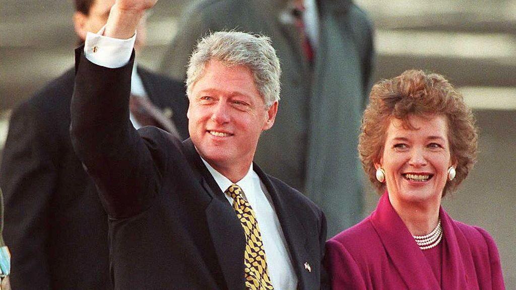 A man with grey hair and black suit waves, beside him a woman with brown hair smiles, she is wearing a pink suit