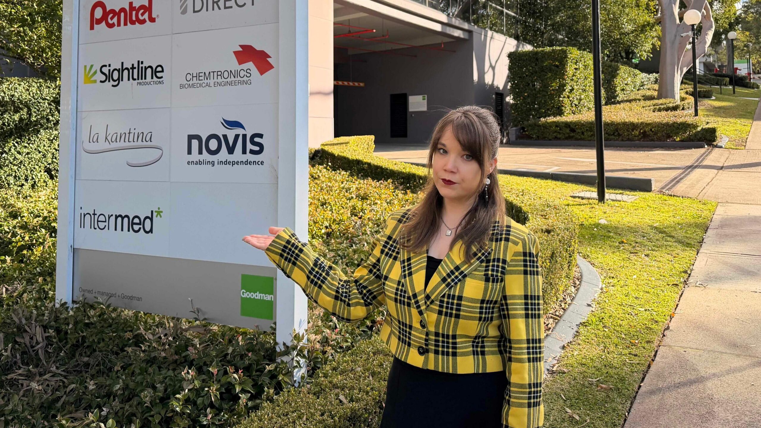 A woman dressed in a yellow and black checked blazer gestures with her hand to a sign in front of a building complex. A green lawn and a gum tree are in the background.