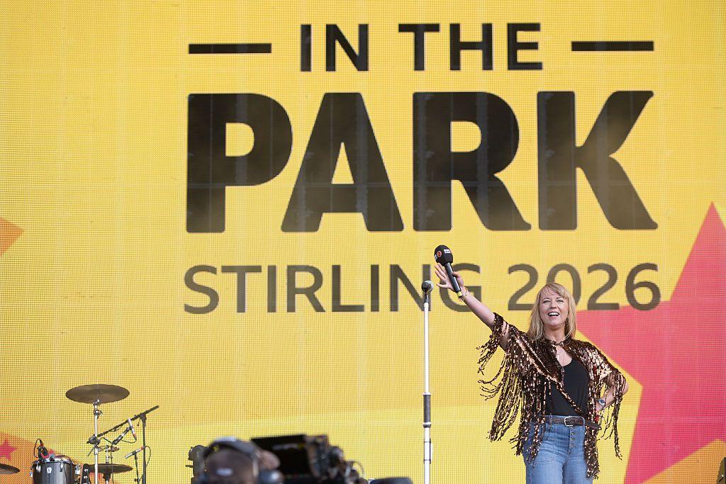 Radio 2 DJ Sara Cox standing on stage holding her microphone aloft. She is wearing blue jeans, a black top and a copper-coloured sparkly tassled jacket