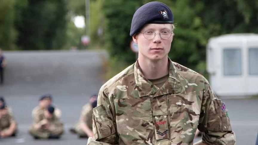 A bespectacled Jackson pictured when he was a cadet. He is wearing a camouflage-patterned uniform and a black military beret. There are other cadets in the background but they cannot be seen very well because the background is blurred. They are all sitting cross-legged on the ground.