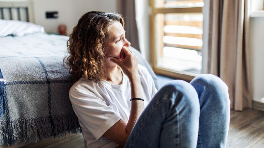 A woman sitting on the floor in her bedroom, looking into the distance.