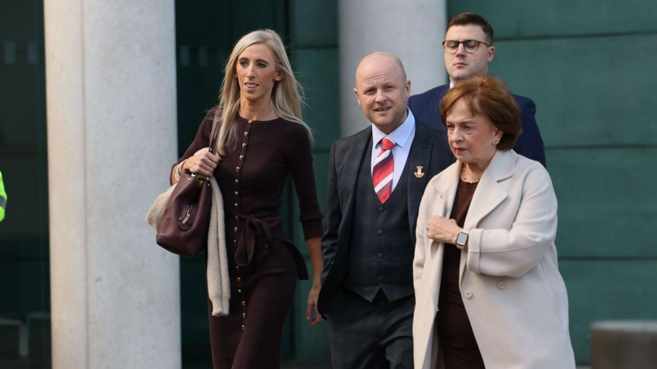 A police man is wearing full uniform walking in front of a group of four people. A blonde woman leads the way wearing a long brown dress. To her left a man is wearing a suit and tie and is bald. Next a woman wearing a long beige coat.