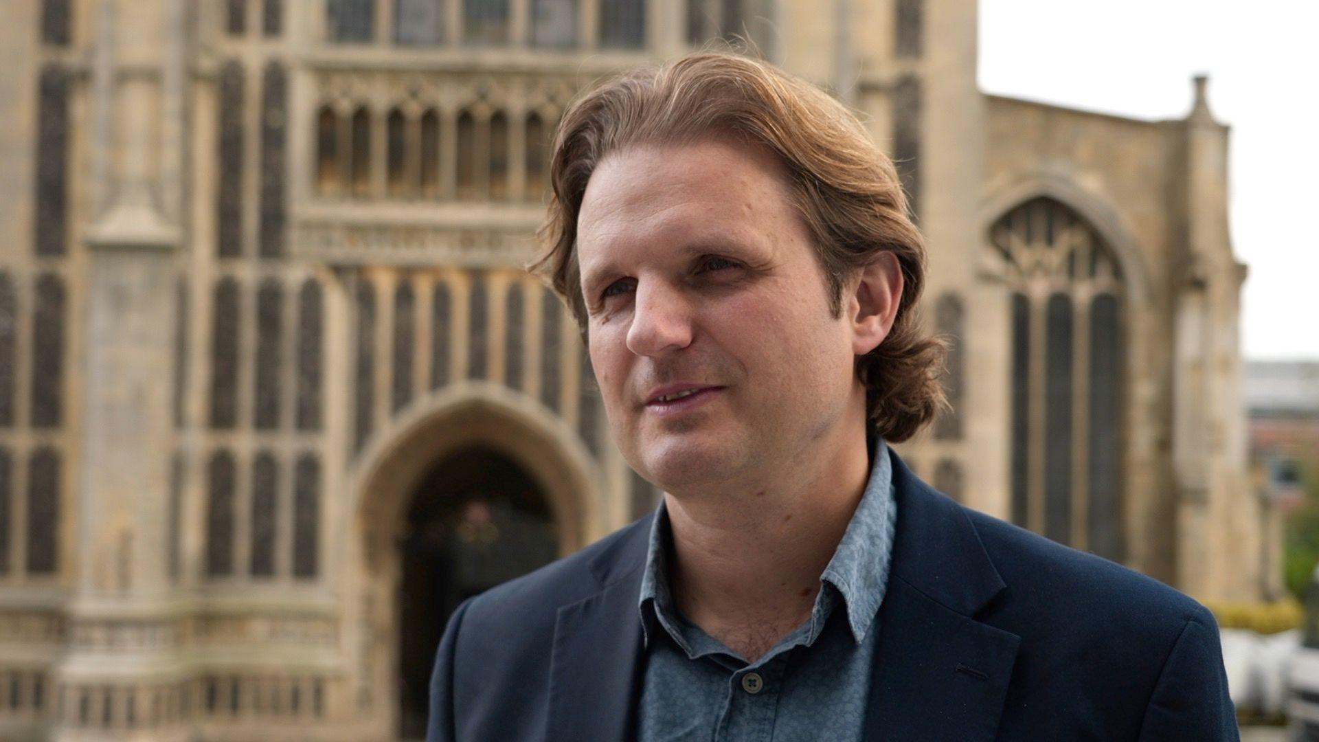 Steffan Aquarone has long, light brown hair. He is standing in front of the St Peter Mancroft church in Norwich and wearing a dark suit.