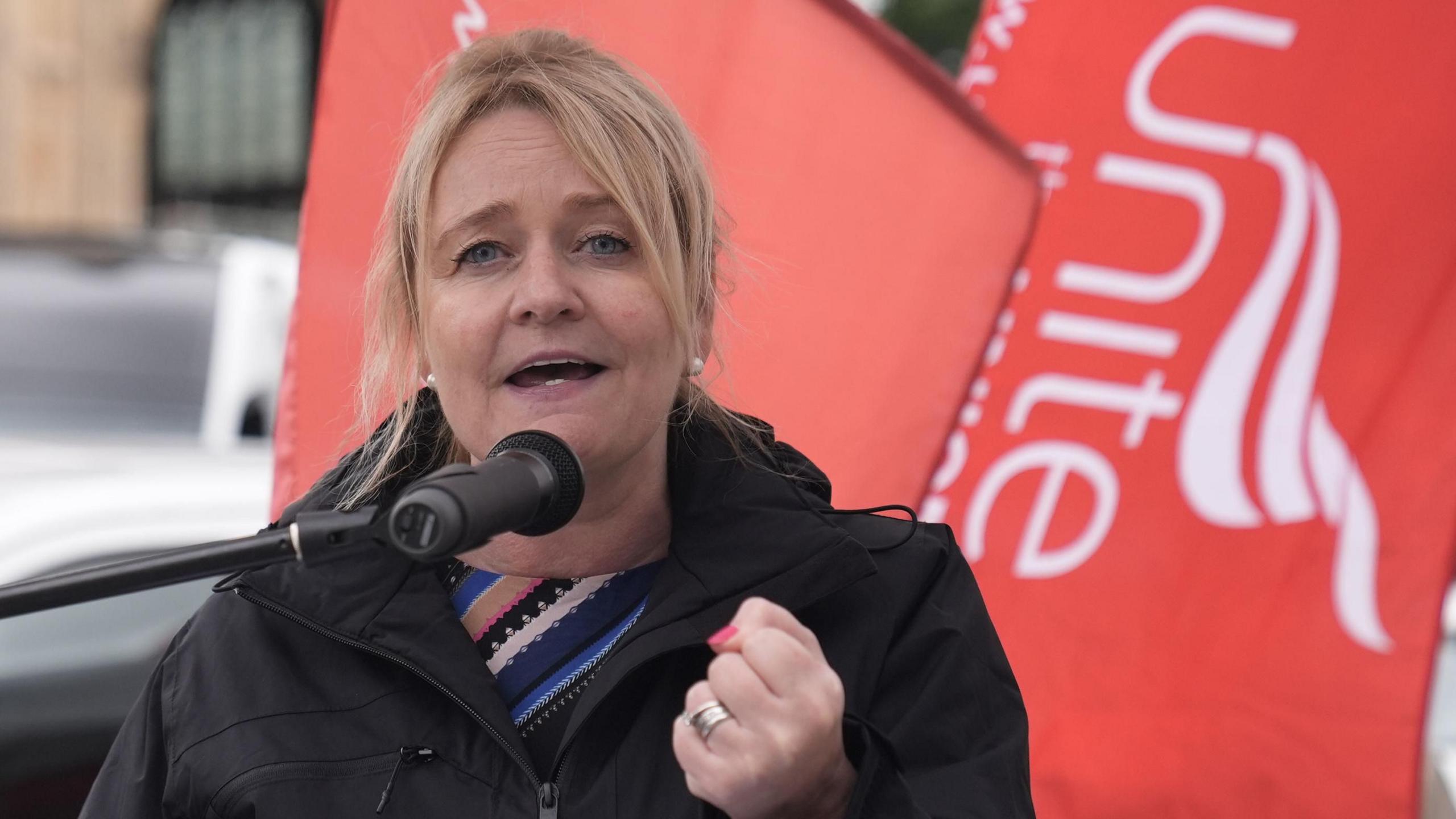Sharon Graham clenches her first as she speaks into a microphone at a rally, with a red Unite flag behind her.