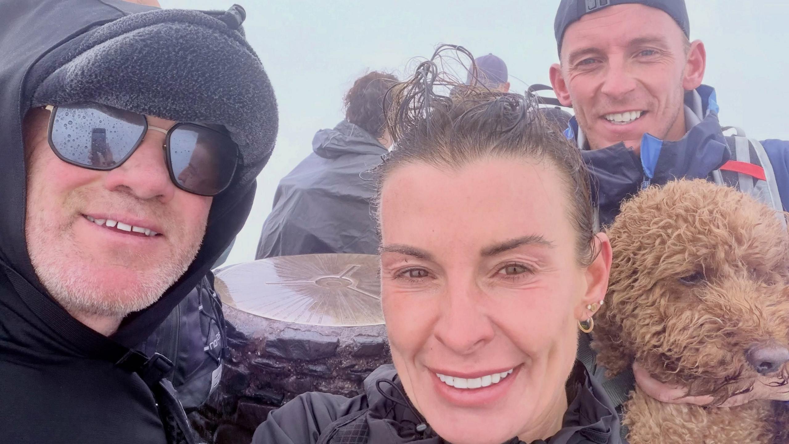 Wayne and Coleen in selfie at the summit in wet and windy weather with hair wet and rain on sunglasses. They are pictured with a friend and a dog behind them.