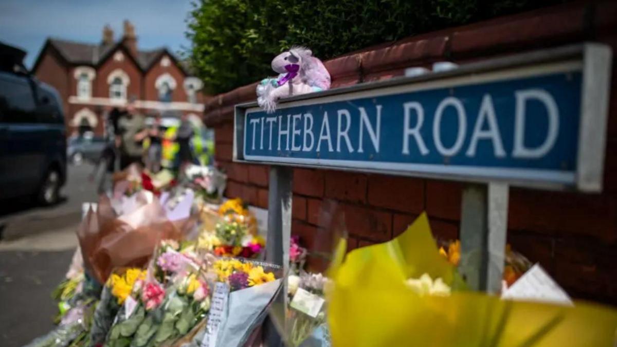 Bunches of flowers and teddies lined up against a wall next to a road sign reading Tithebarn Road.