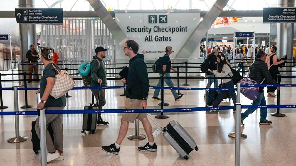 People wearing headphones carry suitcases and walk between aisles in an airport's security line.