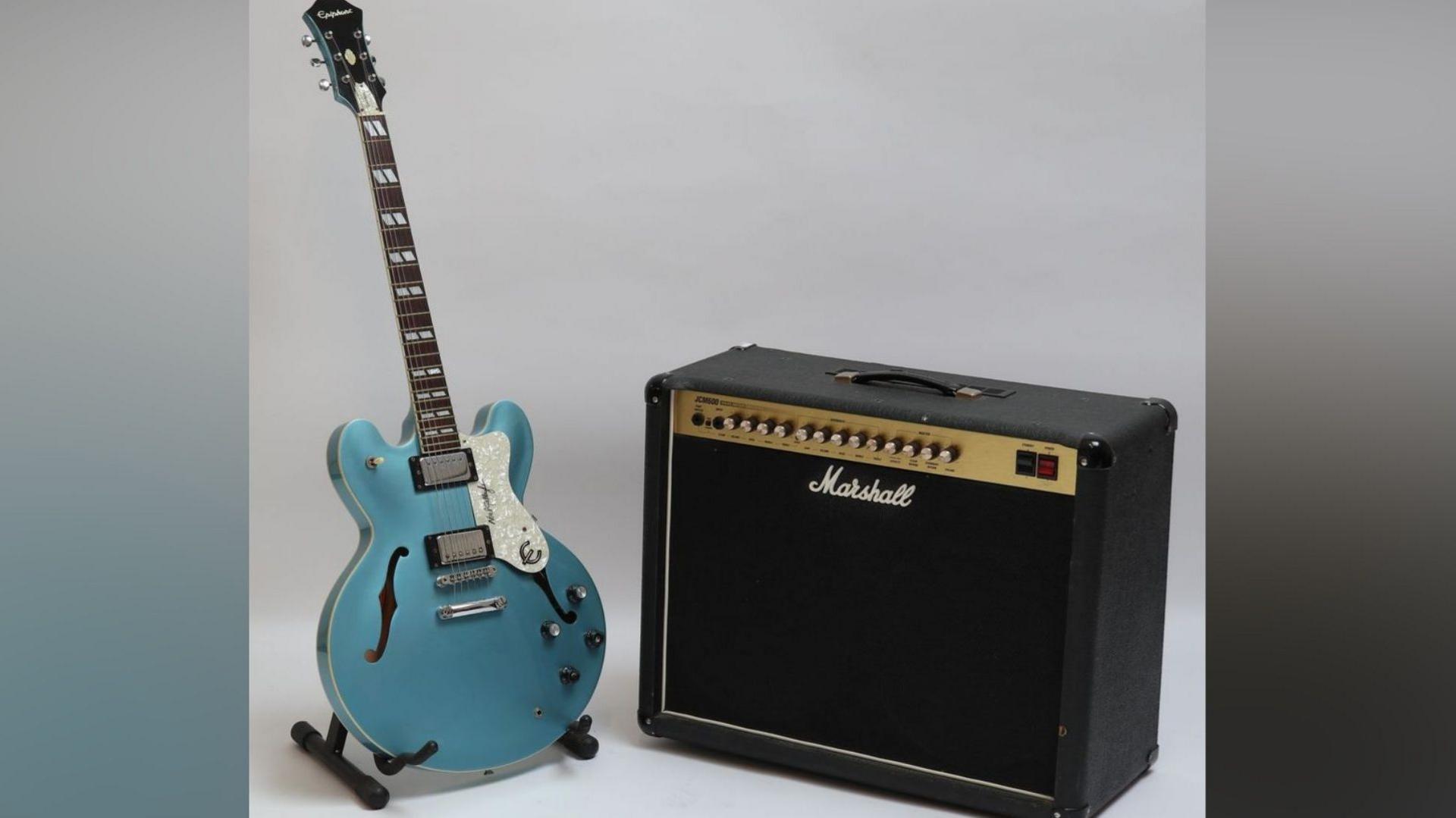 A band-played Epiphone guitar - which is a metallic blue colour. The guitar is resting on a stand next to a Marshall speaker.