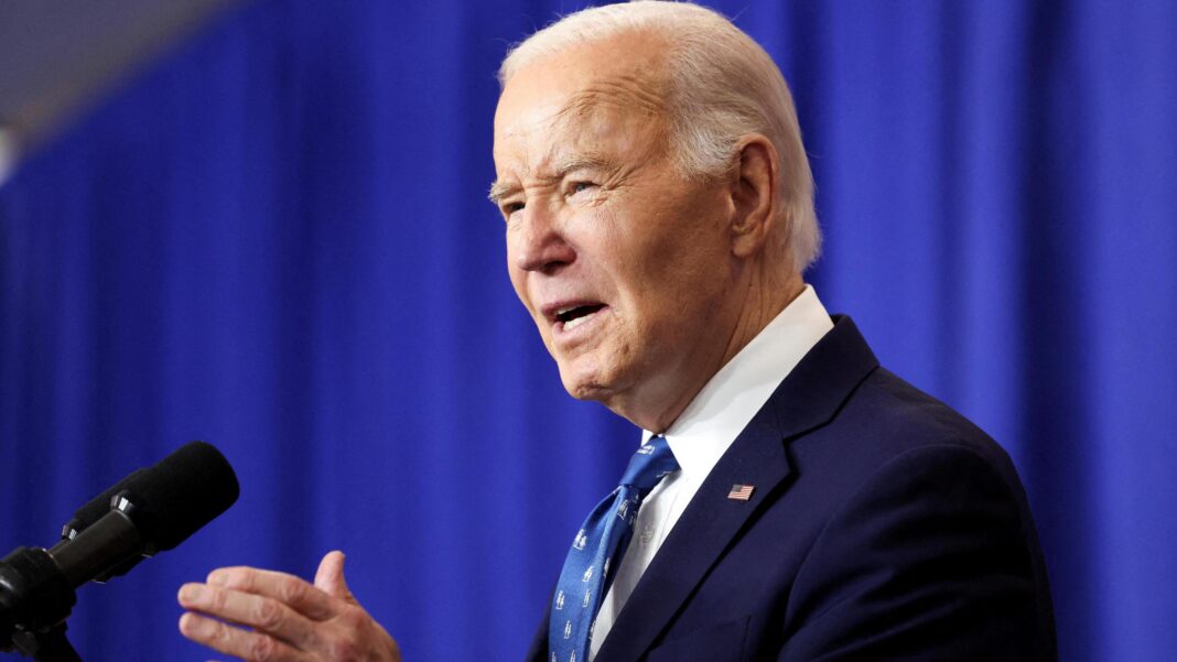 Joe Biden stands side-eye on to the camera, and gesticulates in front of a microphone against a blue curtained backdrop. He wears a navy blazer and white and blue tie.