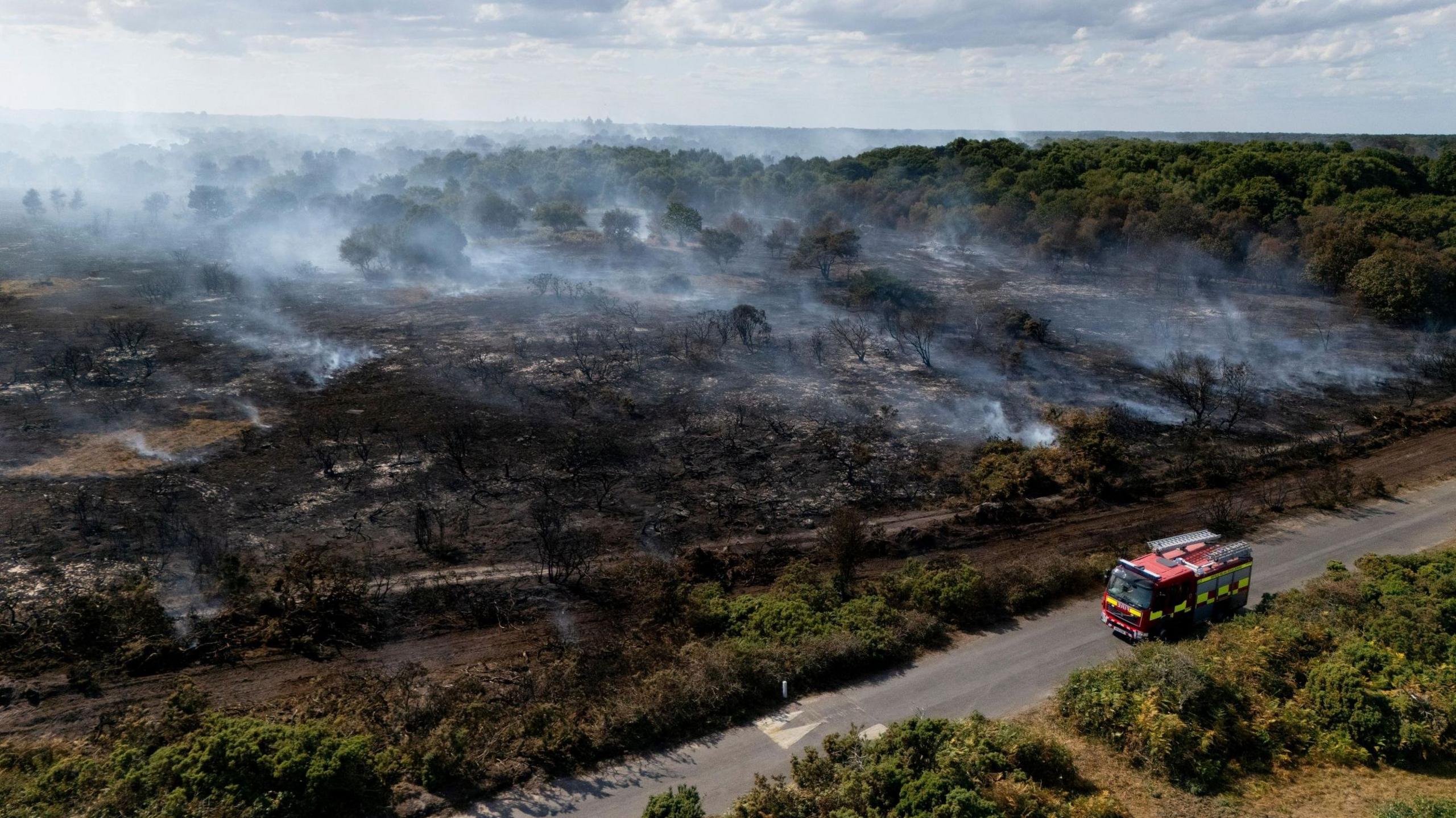 An aerial view taken with a drone shows smoke rising from wildfires near Dunwich Heath in Suffolk. A fire engine can be seen on the road next to a large area of blackened tree stumps
