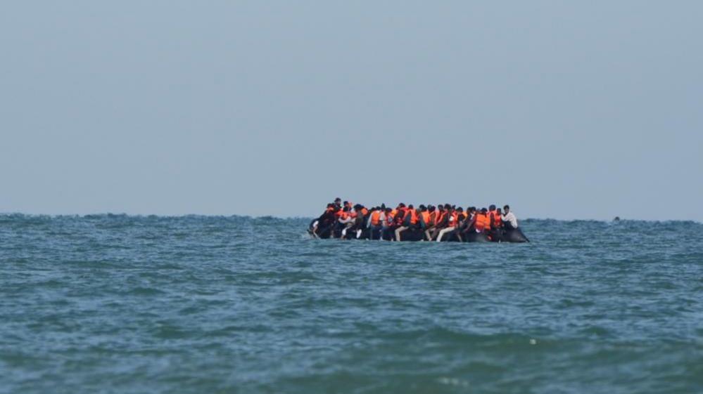 People sit in a dinghy off the coast of Calais in July 2025. Most are wearing orange life jackets.