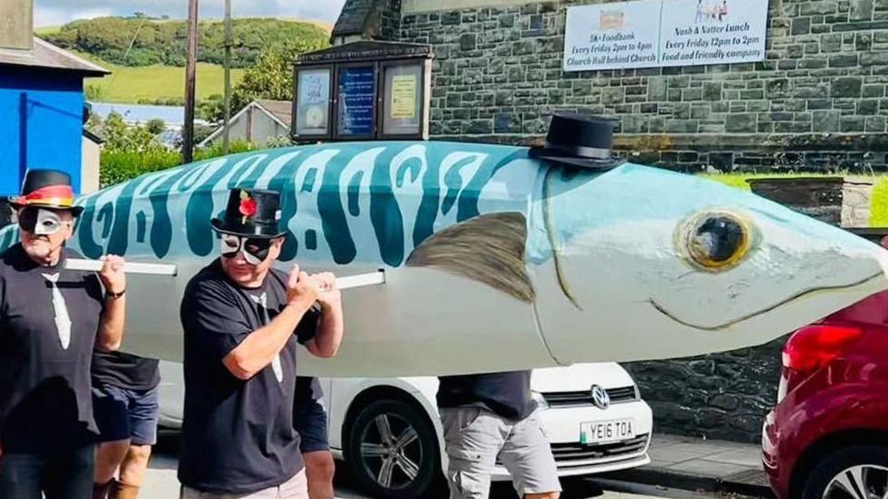 A large blue and white paper mache mackerel. A small black hat is placed on top of the mackerel. It is being carried by six bearers who are wearing black tops with a mackerel printed in the centre. They are wearing black hats, like the one on top of the paper mache mackerel and black and white masks on their faces.