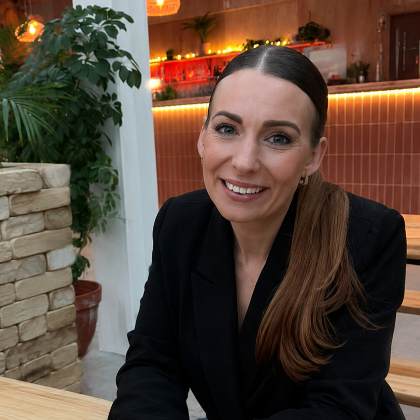 A women with long brown hair in a ponytail, wearing a black jacket, smiles at the camera in front of a bar and a tree