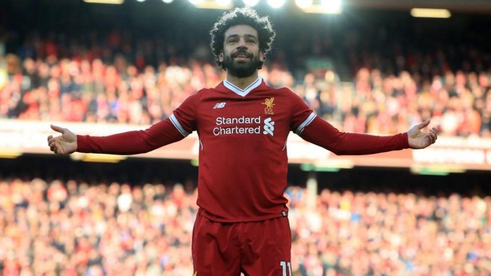 A man with black hair and a beard is wearing a Liverpool kit while standing on the pitch with his arms raised to either side. He is smiling. A large crowd can be seen behind him.
