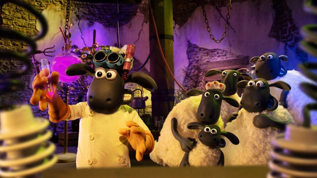 A still from the film showing Shaun the Sheep wearing a lab coat and goggles and holding up a test tube, while a group of other sheep cower in the background