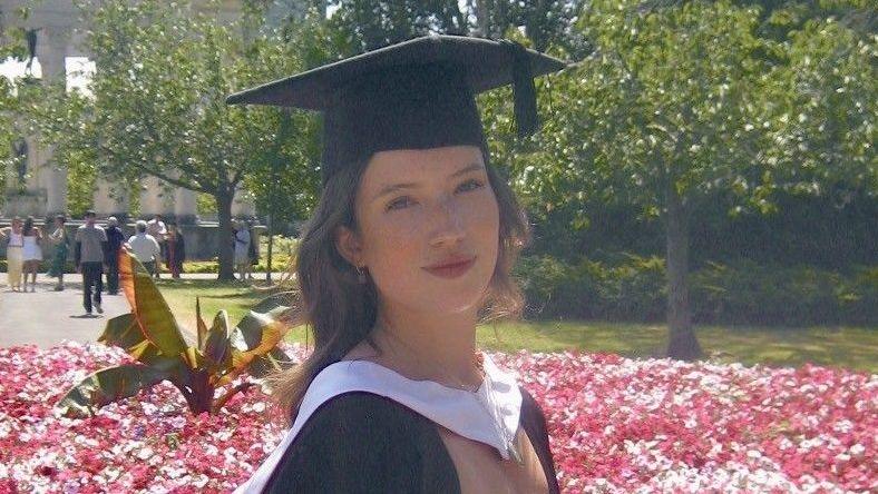 A girl with curly dark hair, wearing a white dress and black graduation cap and gown. She is stood in front of a flowerbed of pink flowers, with trees and people in the background, and is smiling at the camera.
