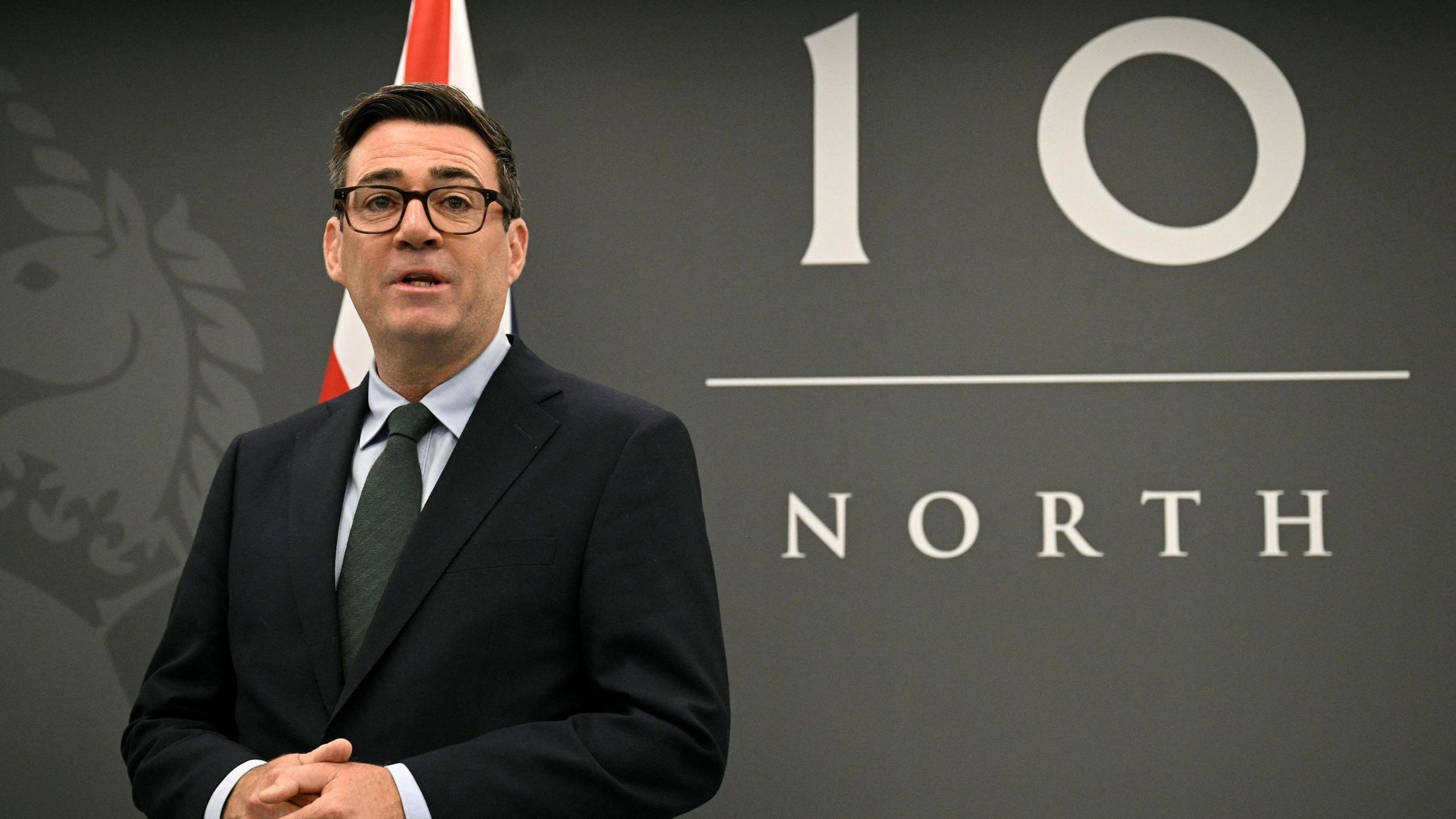 A person in a dark suit and glasses stands in front of a large “10 North” backdrop, with part of a red-and-white flag visible behind them.