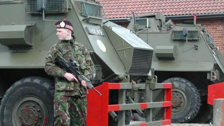 A soldier with a gun. He is stood next to two army vehicles. It appears to be in a residential area.