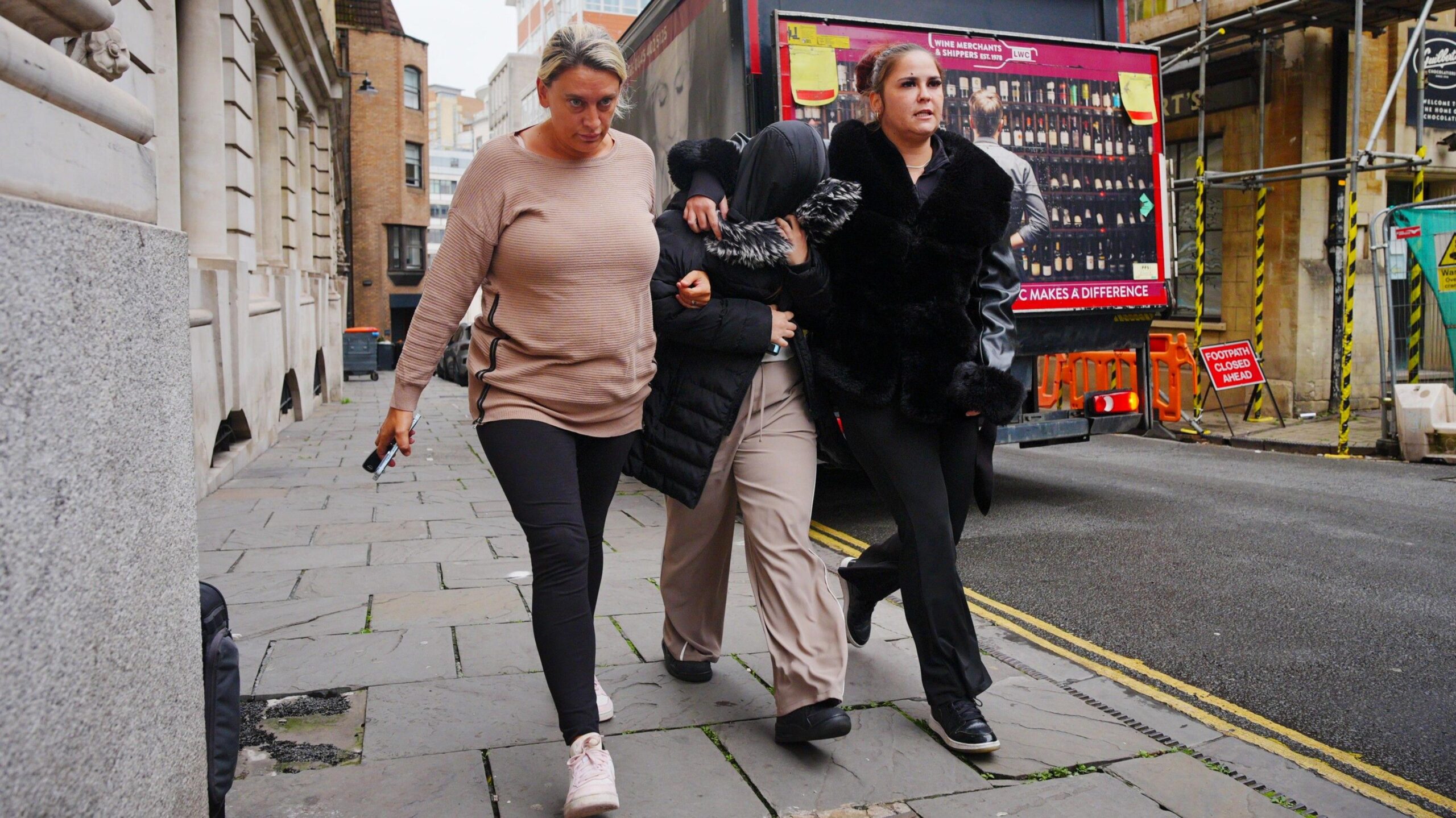 Kelcie Reed wearing light grey trousers and a black puffer coat with a fur lined hood. She is shielding her face from the cameras with her hood as she walks into court. Two women are on either side of her, linking her arm and looking ahead.