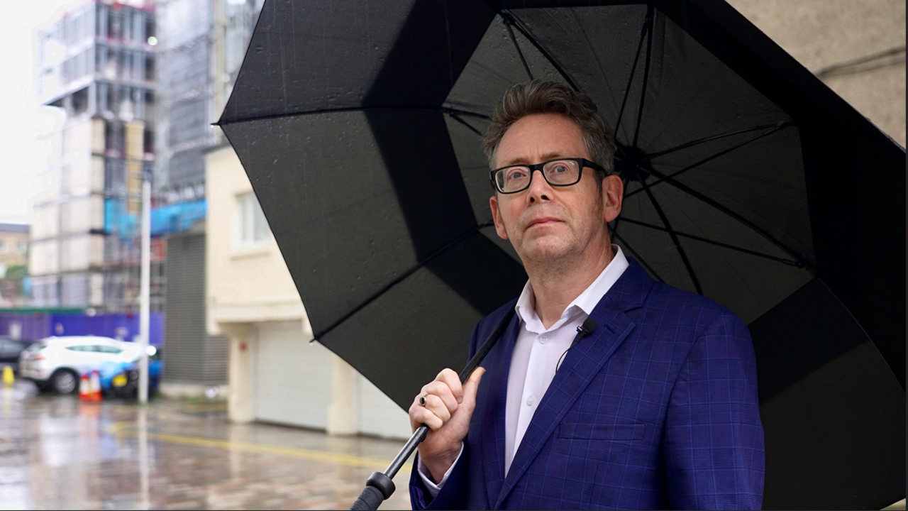 A man, Dr John Boyle, stands on a wet street looking thoughtful. He is wearing a dark blue suit and is holding a large black umbrella.