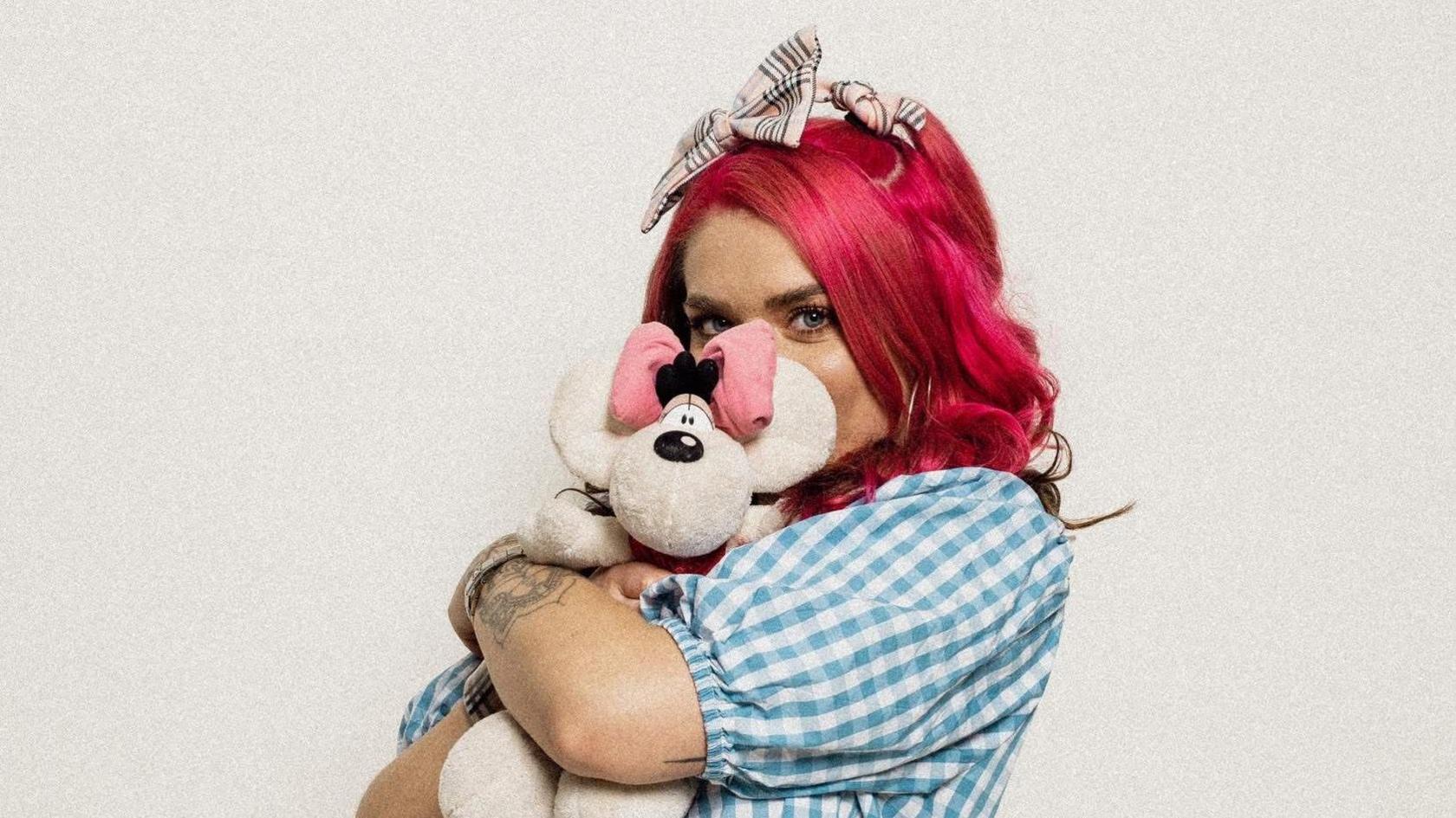 Woman in blue and white checked shirt with dyed pink hair and a tartan hair bow hugging white teddy.