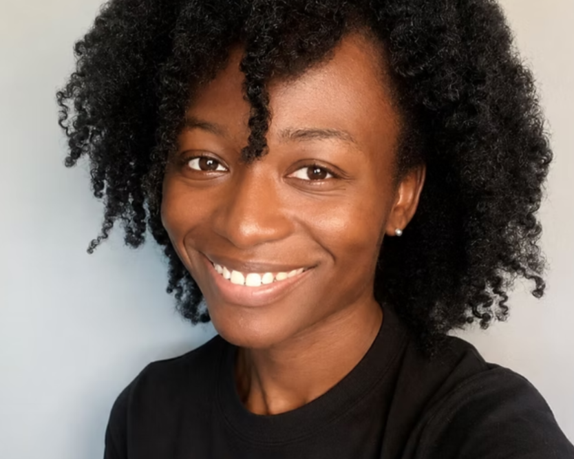 A woman in a black top smiles at the camera, in front of a plain grey wall