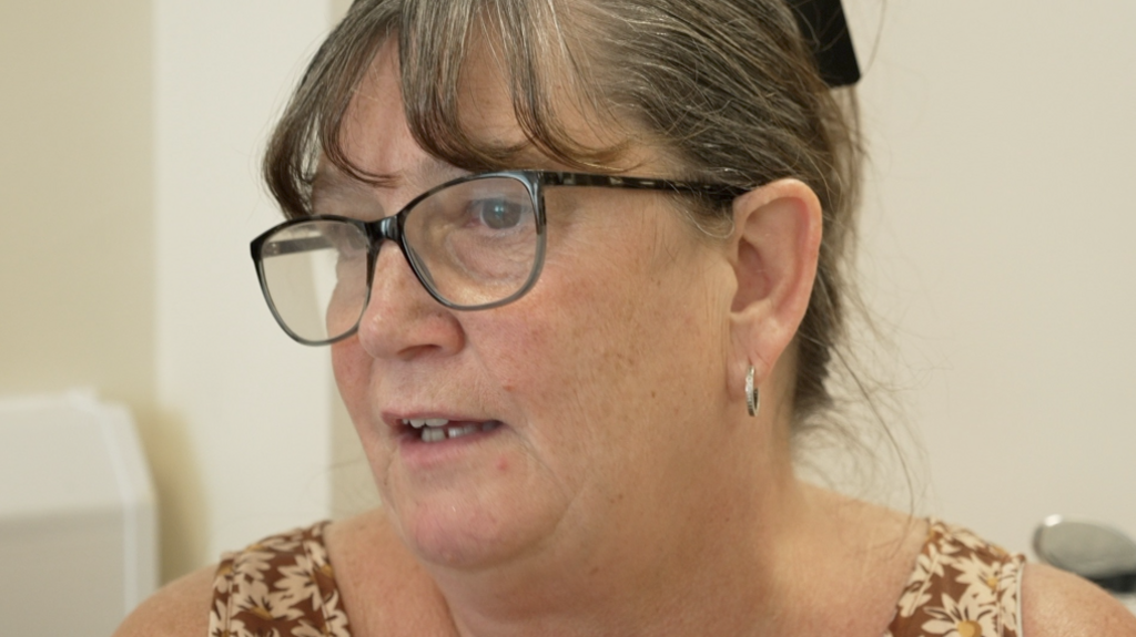 A head and shoulders view of Angela, wearing glasses with tied-back greying hair and hoop earrings