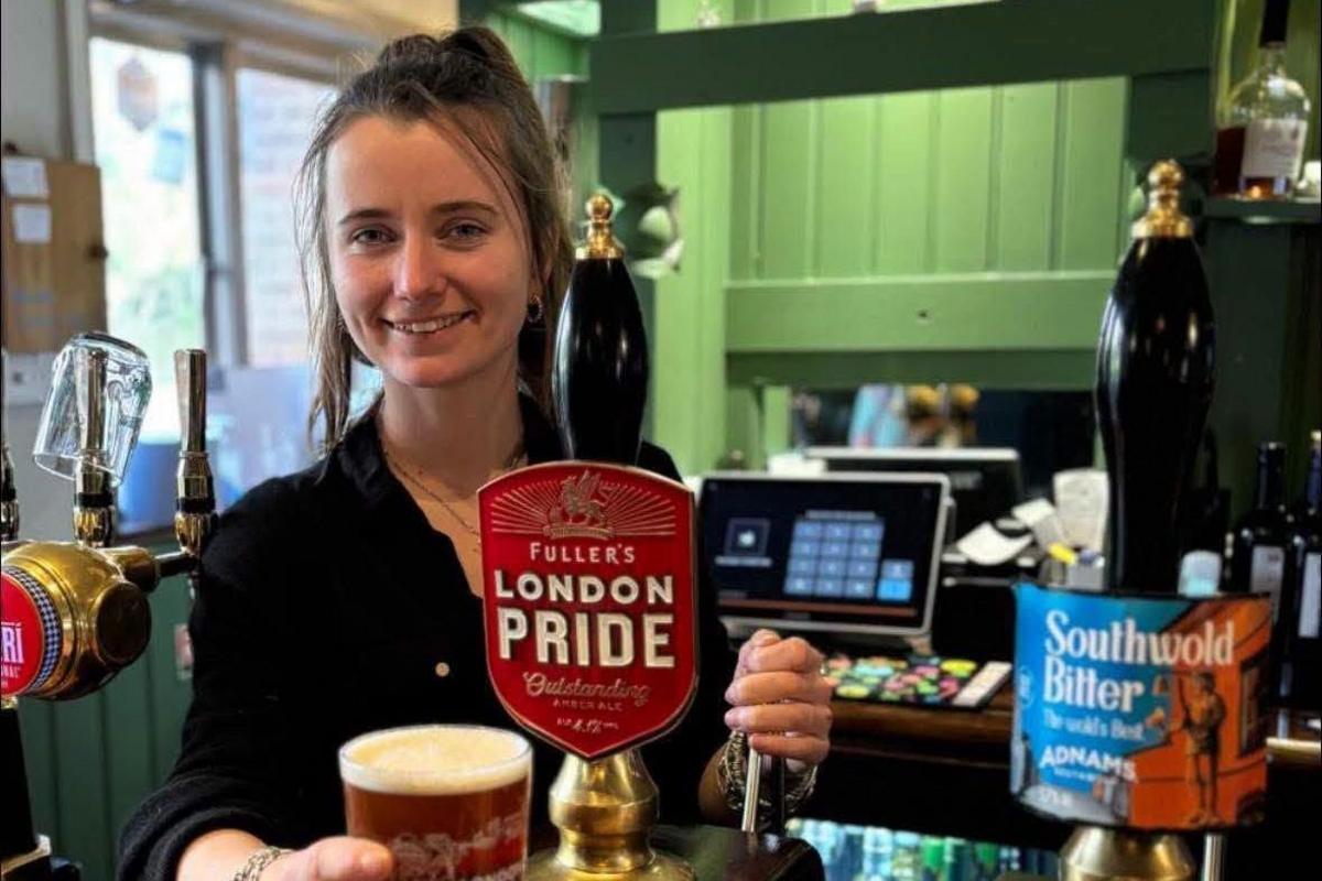 A woman is smiling behind a bar, stood near an ale pump