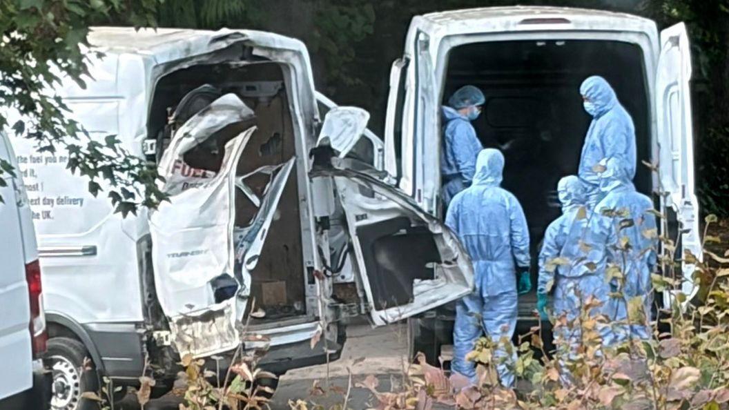 people in blue hazmat suits stand behind a white van next to another white van with the back doors blown off