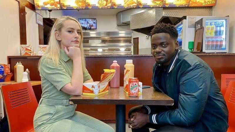 Dimoldenberg and Daniel Kaluuya on Chicken Shop Date. The pair sit opposite each other at a table with boxes of fried chicken in front of them. Dimoldenberg wears a green jump suit while Kaluuya wears a black bomber jacket.