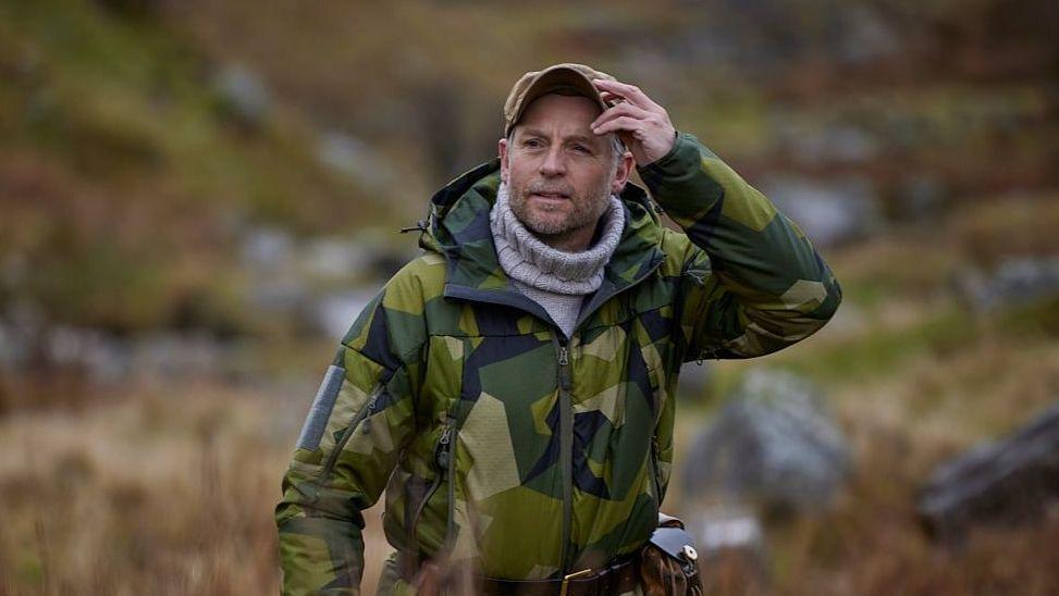 Andrew Price is wearing a green camouflage jacket with a grey turtleneck surrounded by plants and grass.