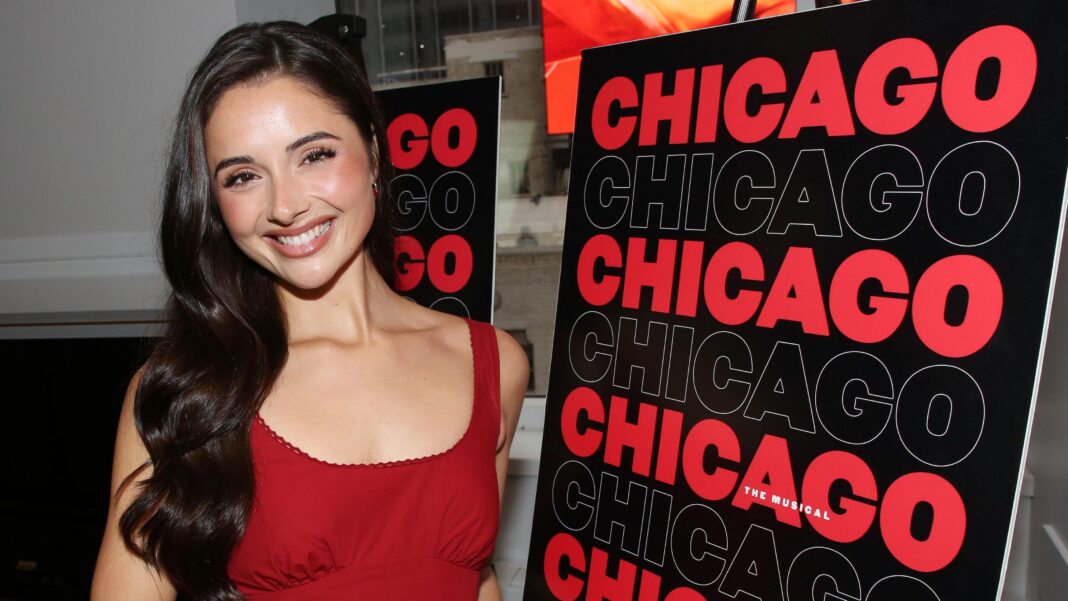 Reece wears a bright red dress and has long brunette hair which sits past her shoulders. She stands in front of a Chicago The Musical sign