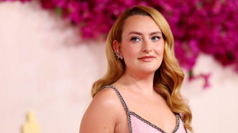 Amelia wears a bejewelled pink dress with straps. She stands against a pink floral background and looks to her side, with her strawberry blonde hair sitting at shoulder length