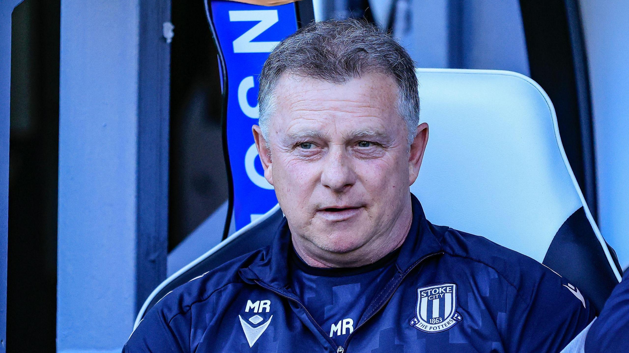 Stoke City boss Mark Robins watches from the dugout. He is wearing a dark blue club tracksuit.