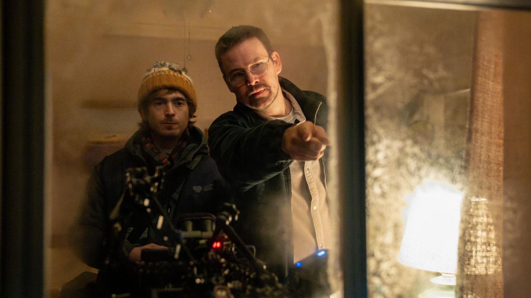 A behind-the-scenes photo from the set of Resident Evil shows Zach Cregger and actor Austin Abrams looking out of a window. Cregger is pointing at something outside, presumably giving instructions to Abrams.