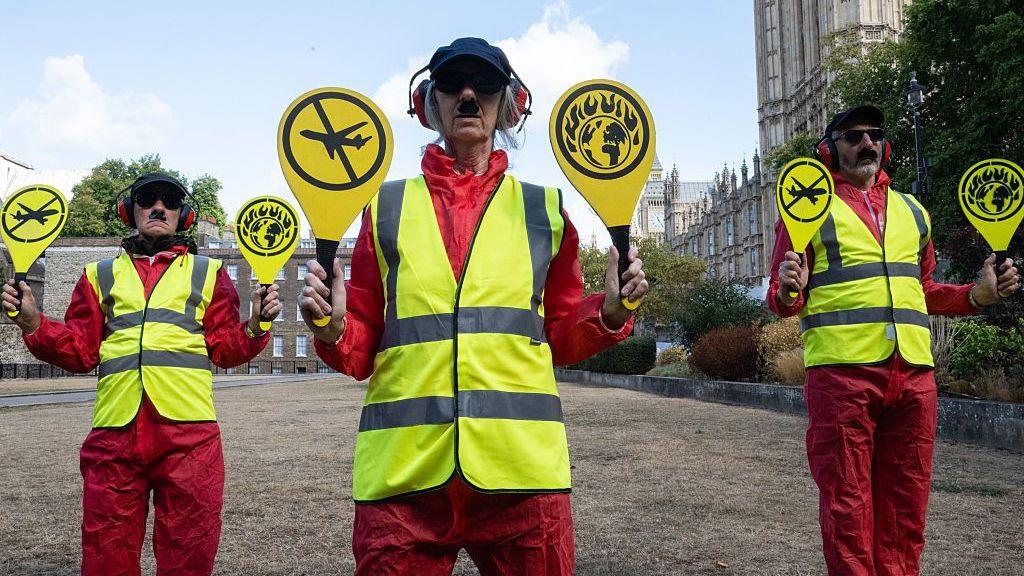 Wearing high vis jackets and holding paddles, three demonstrators mimic runway workers with red overalls, sunglasses and fake moustaches. There is a woman in the middle and two men either side of her. They are stood on a brown lawn to the right the Houses of Parliament are visible, and the sky is blue with a few white clouds.