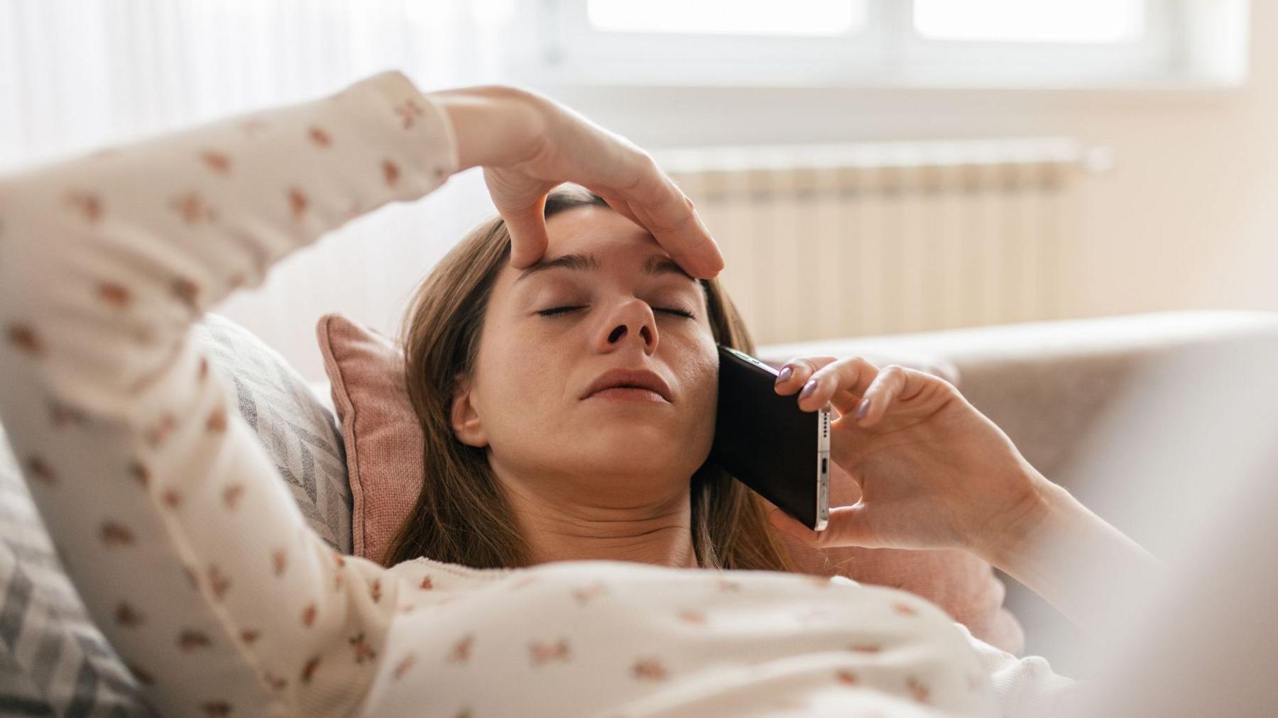 Woman lying back on a sofa with one hand on her forehead and the other hand holding a mobile phone to her ear