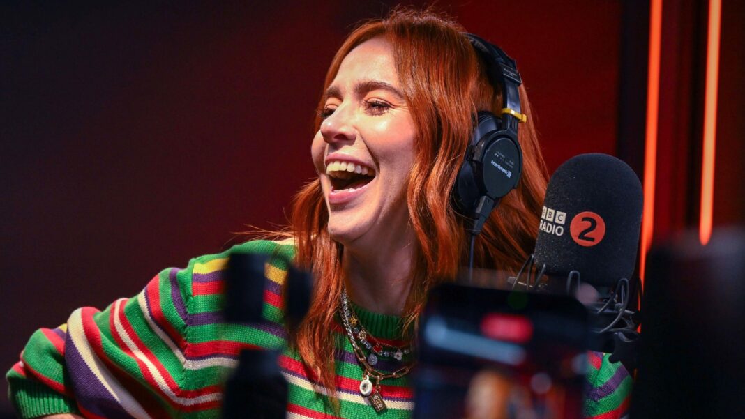 Angela Scanlon takes over Sara Cox’s drivetime slot on Radio 2 Angela Scanlon laughing, wearing headphones in a radio studio front of a microphone with the BBC Radio 2 logo