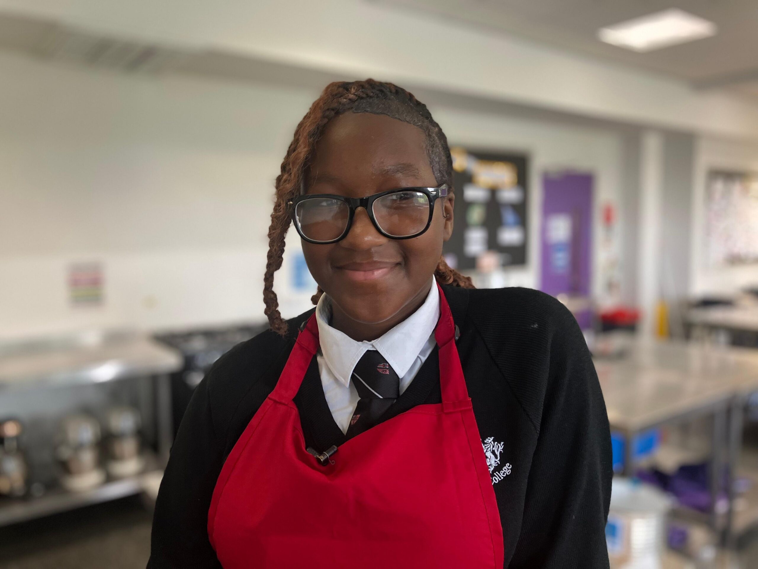 A teenage girl wearing a red apron over her school uniform is smiling at the camera. She's in the middle of a kitchen classroom.
