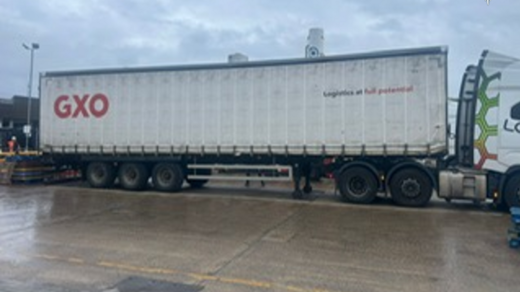Side view of the white tri-axle curtain side trailers with branding for logistics company GXO on an industrial estate. The ground is wet as it has been raining.