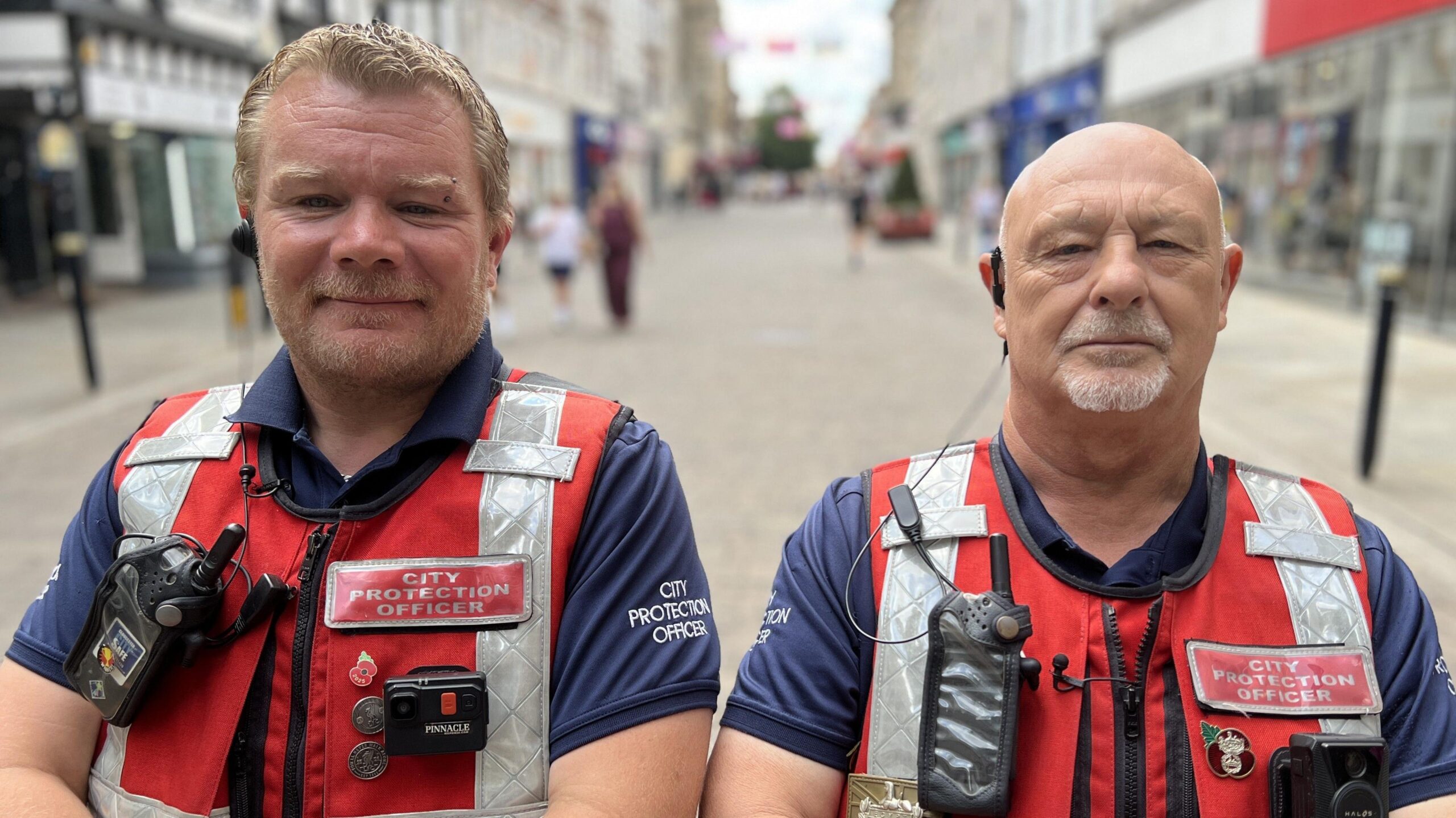 Two men stand with their arms folded facing the camera in the centre of Gloucester. They are wearing red tabards and have radios attached to their uniforms and earpieces in.