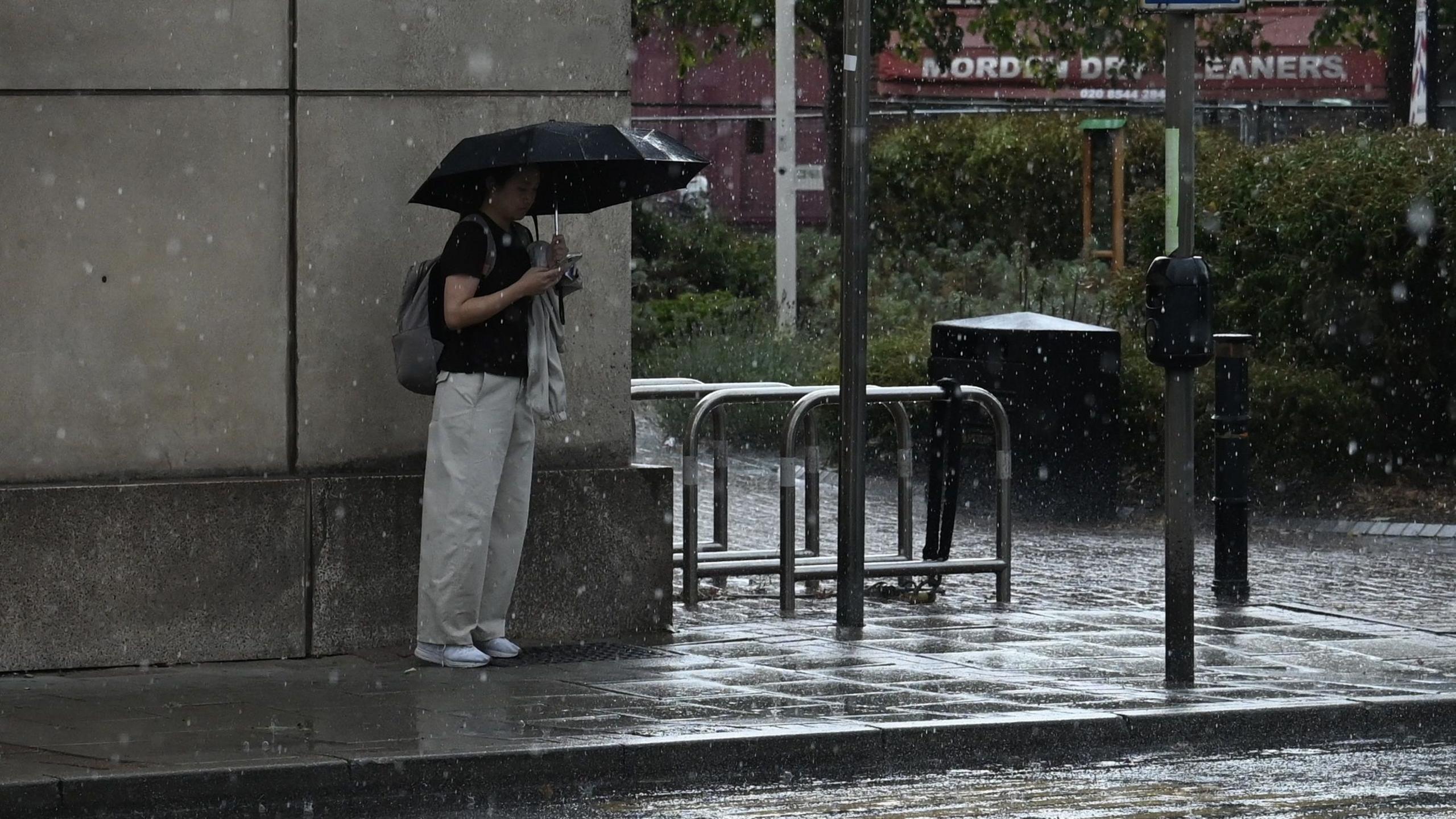 A woman stands with a black umbrella next to a wall in the rain