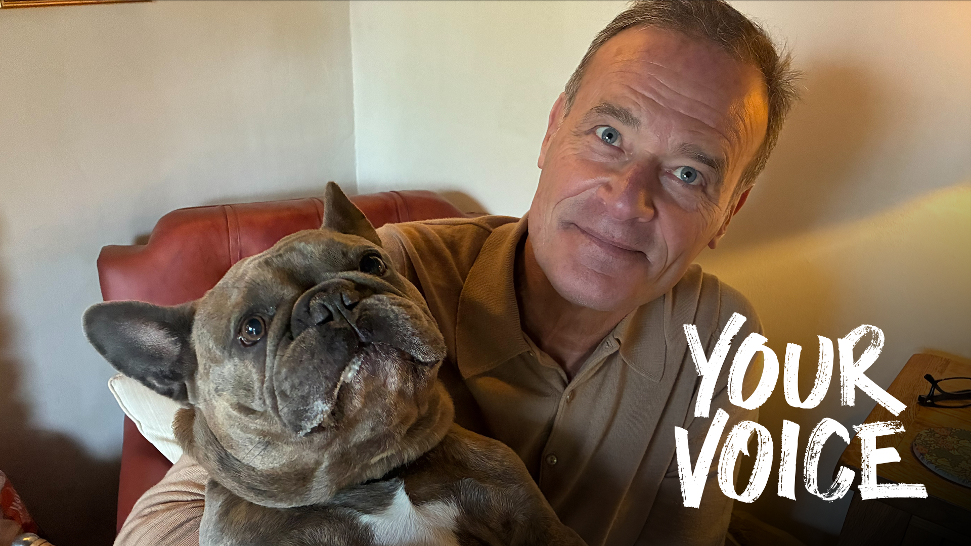 Clive Di Giorgio sitting in his living room in a red leather chair, holding his pug dog Louis. The words YOUR VOICE have been imposed on the bottom right hand corner of the photo.