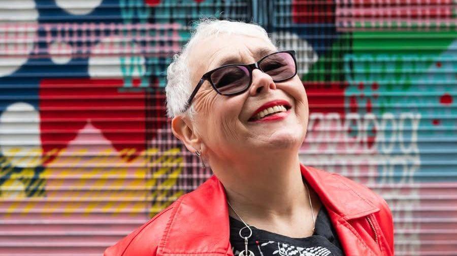 A tightly cropped photo of a woman in her 70s with short grey hair looking up at the sky, smiling. She has sunglasses on, and bright red lipstick that matches her leather jacket. Graffiti is in the background, slightly out of focus.