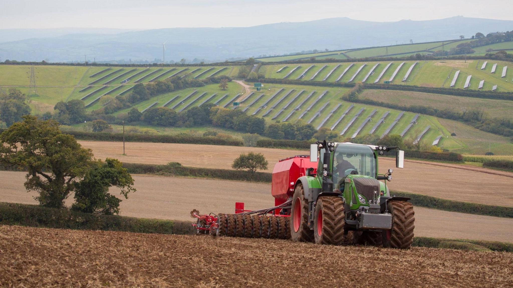A large green tractor is pulling a red cultivator and drill along a ploughed field. In the background, solar panels are spread over a field on the hillside.